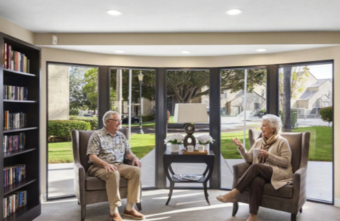 A group of elderly people are sitting around a table, smiling and enjoying their time together. They are holding green objects in their hands, possibly flowers or plants. The room is decorated with potted plants and vases, creating a pleasant atmosphere for the group. AI generated content